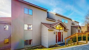 En bygning med flere etager, en garage, et mindre tilbygget hus med en rød dør, og en rampe af træ der fører til indgangen.