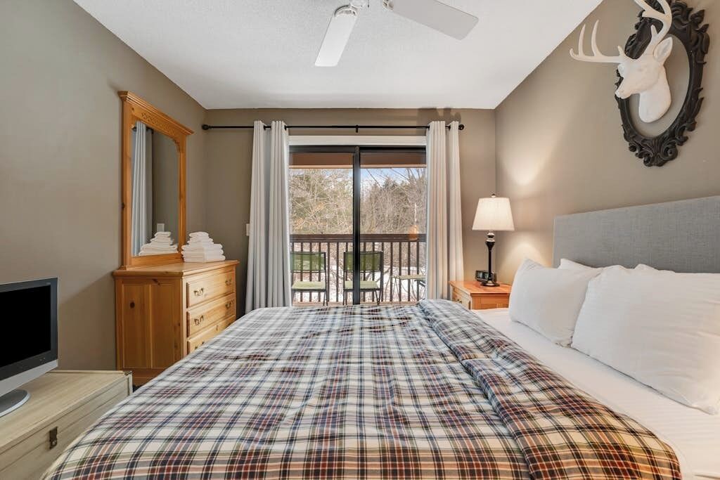 A bedroom with a bed, a dresser, a television, a ceiling fan, a balcony with a view, and a deer head decoration.