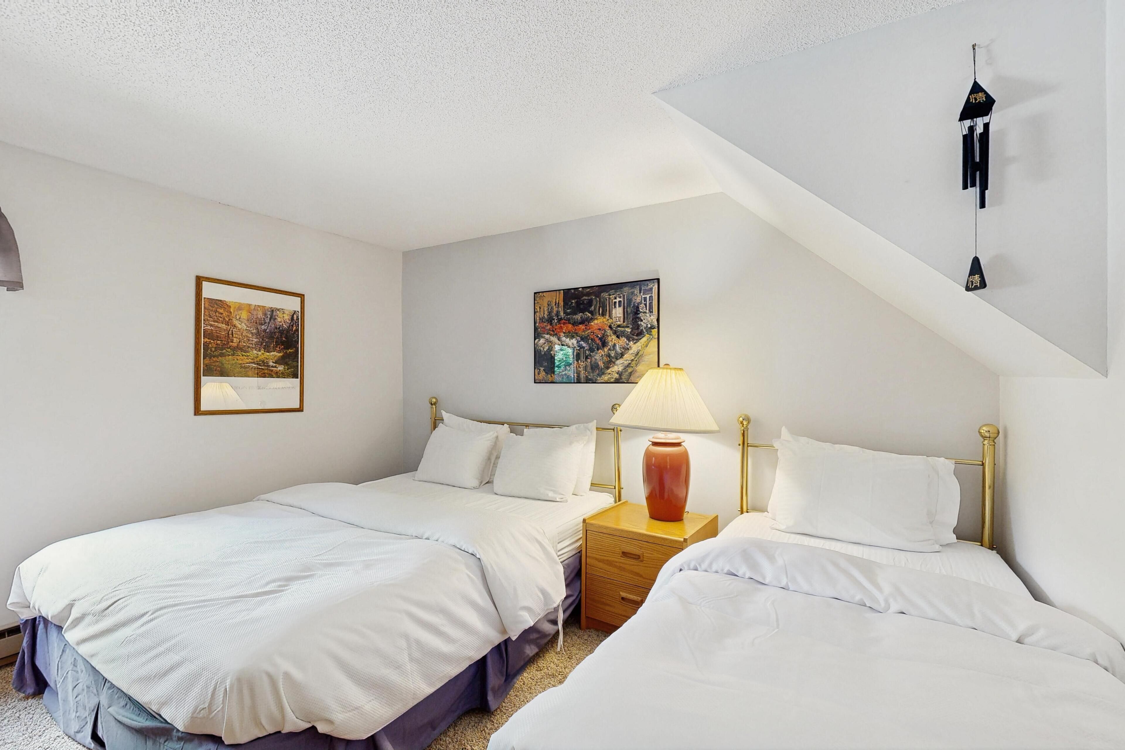 A hotel room with two beds, a nightstand with a lamp, and two wall-mounted paintings.