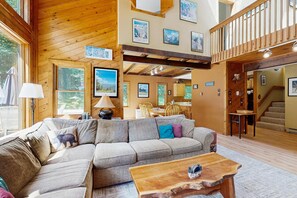 Ein gemütliches Blockhaus-Interieur mit einer großen Holzbalkendecke, einer Eckcouch, einer Essecke und einer Treppe, die zu einer oberen Ebene führt.
