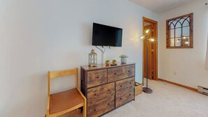 A room with a wooden chair, a wooden dresser with drawers, a flat-screen TV mounted on the wall, a floor lamp, a window with a decorative frame, and a door leading to another room. - Cozy retreat near slopes (Dover)