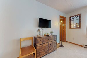 A room with a wooden chair, a wooden dresser with drawers, a flat-screen TV mounted on the wall, a floor lamp, a window with a decorative frame, and a door leading to another room. - Cozy retreat near slopes (Dover)