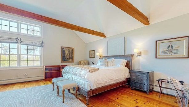A bedroom with a large bed, wooden beams, a window, a dresser, and artwork on the walls.