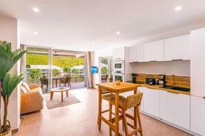 A modern kitchen with white cabinets, a wooden dining table, and a balcony with a view of a green hillside. - Prestigious Serenity Bay beachfront suite (Anse Marcel)
