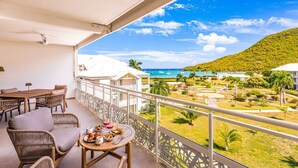 A balcony with wicker furniture, a table with food, and a view of a resort with buildings and a beach. - Serenity Bay – Prestige Suite Anse Marcel beach front (Anse Marcel)