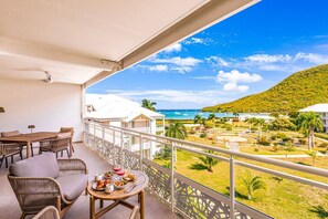 A balcony with wicker furniture, a table with food, and a view of a resort with buildings and a beach. - Prestigious Serenity Bay beachfront suite (Anse Marcel)