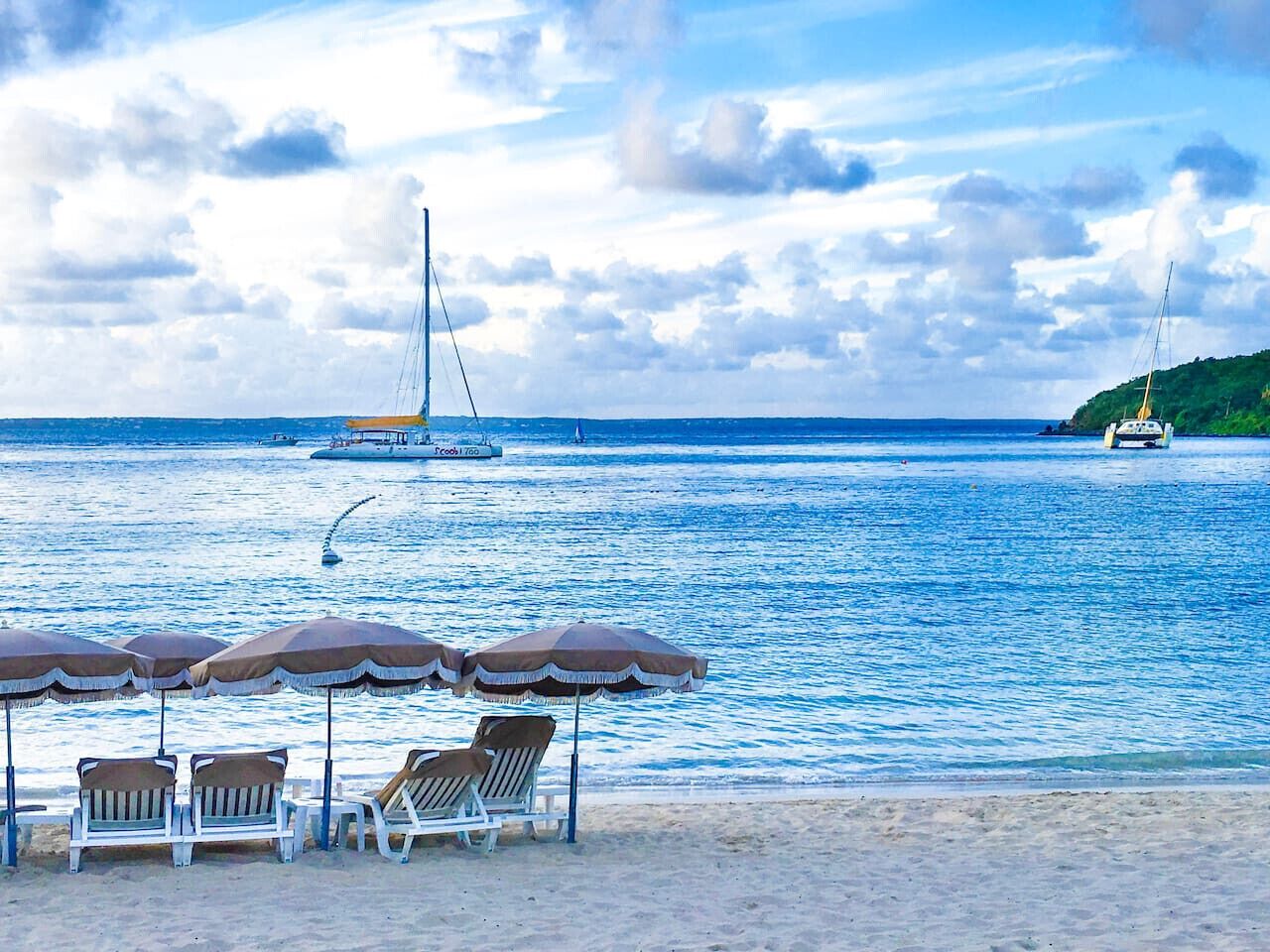 Une plage avec des chaises longues et des parasols, un voilier et une petite île au loin.