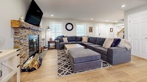 Una sala de estar con chimenea de piedra, un sofá modular grande, un televisor de pantalla plana y una escalera que sube a otro nivel.