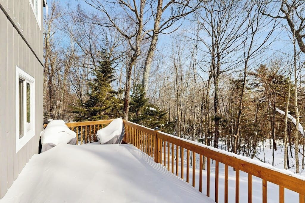 A wooden deck with snow-covered railings, a building with windows, and a forested background.