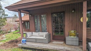 Una terraza de madera con un sofá y una silla de mimbre, un tanque de propano y un ventanal grande.