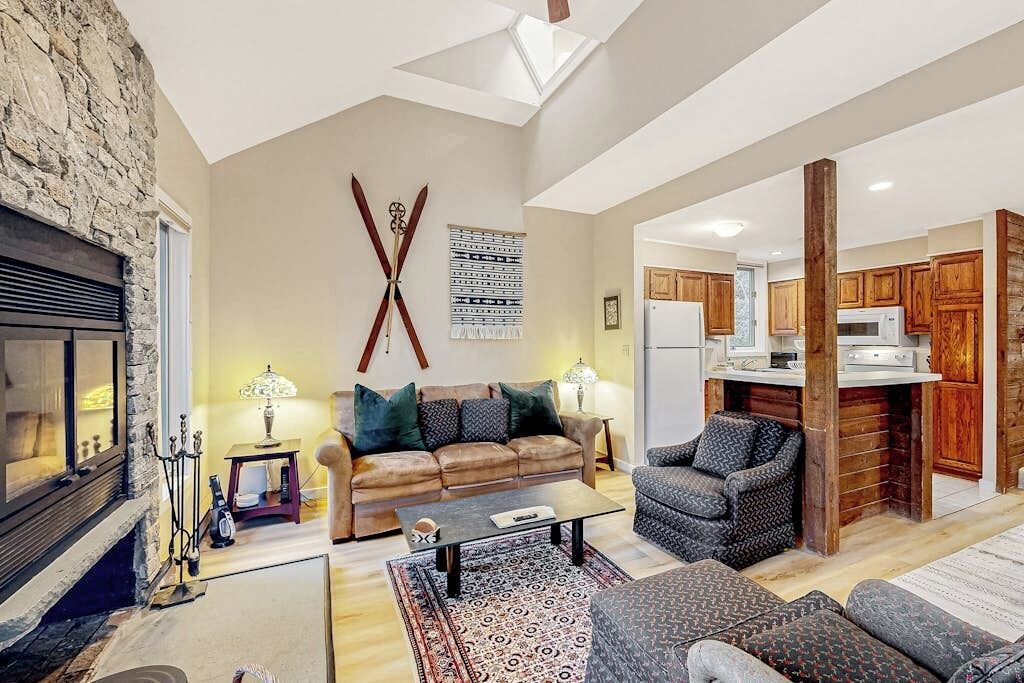 A spacious living area with a stone fireplace, a large sofa, a coffee table, and a kitchen with wooden cabinets.