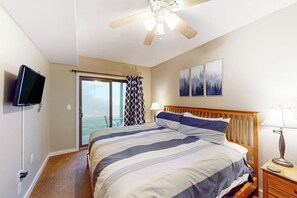 Ein Schlafzimmer mit einem Bett, Deckenventilator, Fernseher, einem Fenster mit Vorhängen und einem Bild an der Wand.