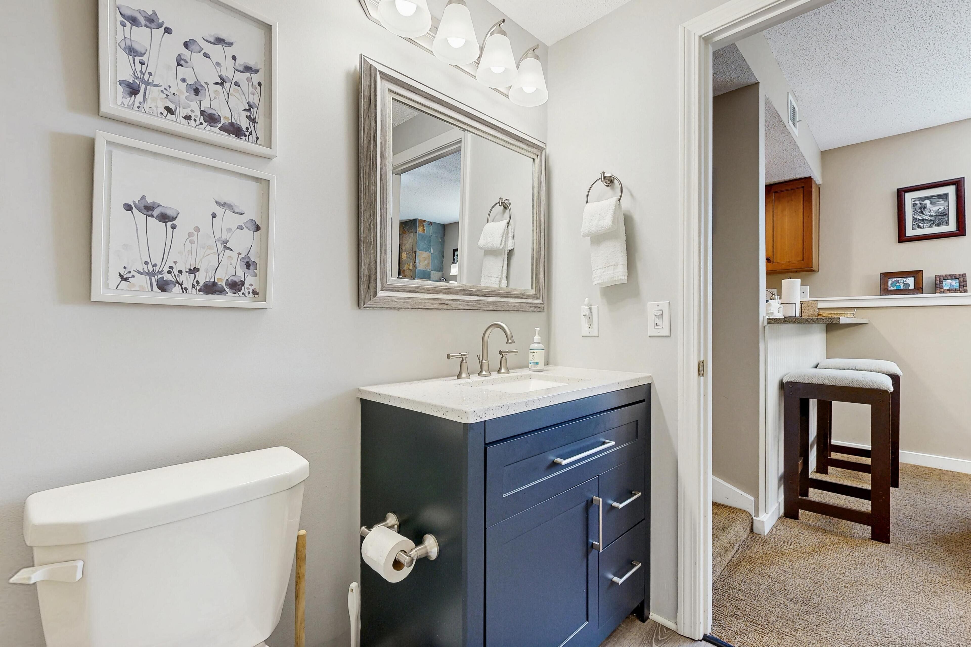 A bathroom with a toilet, sink, and mirror. There are framed pictures on the wall and a towel rack.