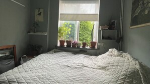 A bedroom with a large bed, a window with blinds, a hanging light fixture, and a shelf with potted plants. - Cozy flat in a quiet, leafy neighborhood in central Malmö (Malmö)