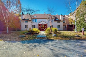 A two-story house with a central red door, a front porch, and a driveway leading to the entrance. - Walk-to-shuttle condo, balcony, fireplace, jacuzzi, pool, hot tubs, sauna (Dover)