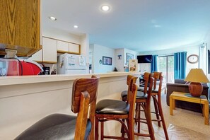 A modern kitchen with wooden cabinets, a dining area with wooden chairs, a sofa, and a lamp. - Walk-to-shuttle condo, balcony, fireplace, jacuzzi, pool, hot tubs, sauna (Dover)