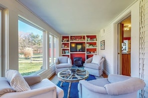 A living room with a fireplace, a bookshelf, and a large window overlooking a yard. - Cozy dog-friendly home, fenced yard, gas fireplace, BBQ, wifi, partial a/c, w/d (Dorset)