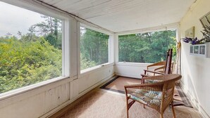 A covered porch with a view of trees, a wicker chair, and a small table. - Central home: near town, lake, skiing, rail trial, snowshoe from your door (Wolfeboro)