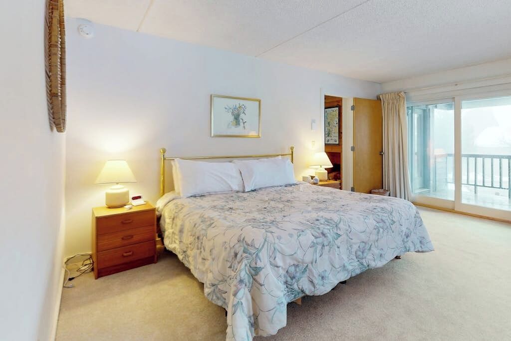 A bedroom with a bed, bedside tables, a nightstand, a window with curtains, and a door leading to another room.