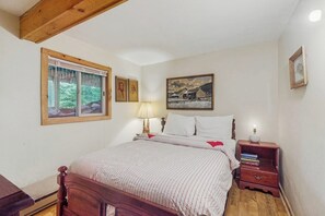 Ein Schlafzimmer mit einem Holzbett, einem Nachttisch mit Büchern, einem Fenster mit Vorhängen und einem Wandbild.