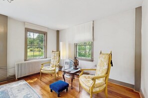 A room with two upholstered chairs, a small table, a radiator, and a window with a view of greenery. - Historic Vermont farmhouse in 100+ acres, balcony, sunroom, office, window a/c (Chester)
