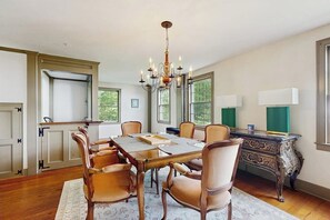 Une salle à manger avec une grande table, des chaises, un lustre et un bureau avec une lampe.
