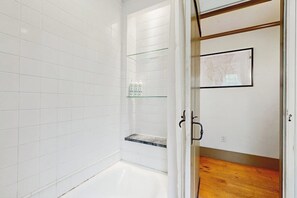 Une salle de bain aux murs recouverts de carreaux blancs, une étagère en verre où sont posés des articles de toilette, un plancher en bois et une carte encadrée accrochée au mur.