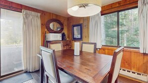 Une salle à manger aux murs lambrés, avec une grande table, des chaises, un lustre et une vue sur la forêt grâce à de grandes fenêtres.