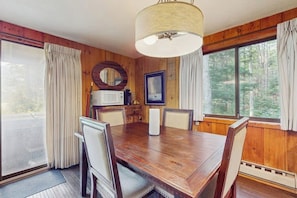 Ein Esszimmer mit Holzwandverkleidung, einem großen Tisch, Stühlen, einer Kronleuchter und Blick auf den Wald durch große Fenster.