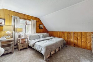 Ein Schlafzimmer mit schrägverdeckter Decke, Holzwänden, einem Bett mit grauer Bettdecke, einem Nachttisch mit Lampe und einem Fenster mit Vorhängen.