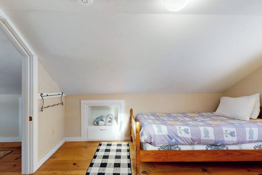 A bedroom with a wooden bed, a checkered rug, a white door, and a small white shelf with a cushion.