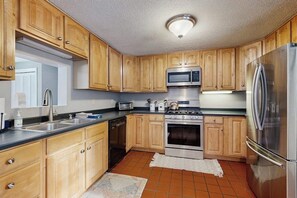 A kitchen with wooden cabinets, stainless steel appliances, a tiled floor, and a window with blinds. - White Mountain home w/ yard near skiing, hiking & tubing (Campton)