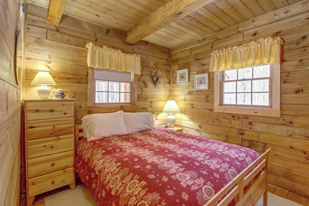 A wooden cabin bedroom with a bed, two bedside tables, a dresser, and two windows with curtains.