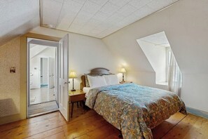 A bedroom with a sloped ceiling, a bed with a patterned bedspread, two bedside lamps, and a window with sheer curtains. - Charming home close to skiing & golf, wifi, piano, w/d, soaker tub (Ludlow)
