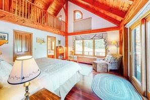 A spacious bedroom with a large bed, a sitting area with a sofa and armchair, a wooden staircase, and large windows. - Mountain-view house with hot tub, AC & amazing views, near Okemo Resort & skiing (Ludlow)