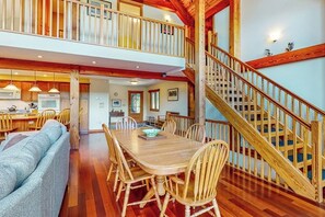 A spacious dining area with a wooden table and chairs, a sofa, and a staircase. - Mountain-view house with hot tub, AC & amazing views, near Okemo Resort & skiing (Ludlow)
