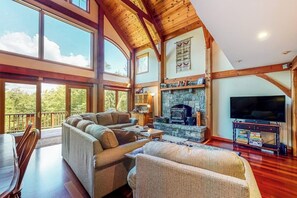 A spacious living room with a stone fireplace, a television, and a balcony with a wooden railing. - Mountain-view house with hot tub, AC & amazing views, near Okemo Resort & skiing (Ludlow)