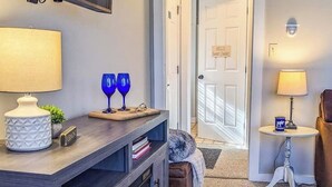 Una habitación con una consola de madera oscura, una lámpara blanca, dos jarrones de vidrio azul, una planta pequeña y una puerta con un letrero que dice 'HELLO Sweet Treats!'.