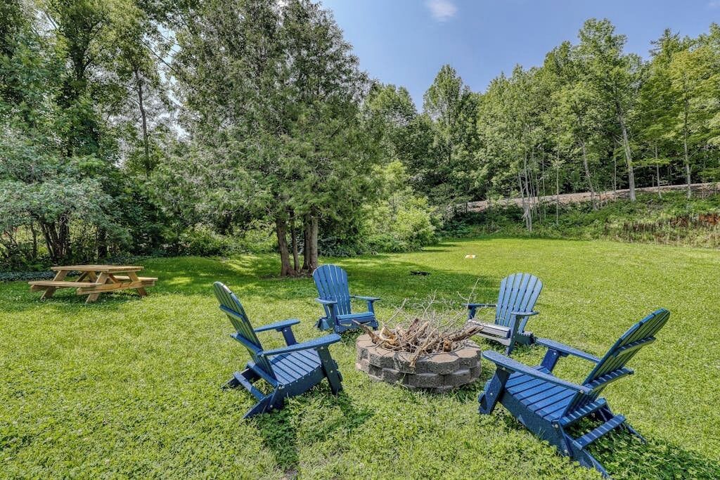 Un patio trasero con un brasero, cuatro sillas tipo Adirondack y una mesa de picnic rodeado de árboles.