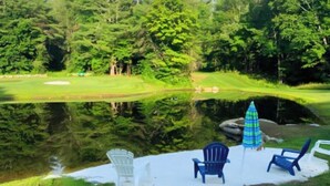 A serene lakeside setting with a golf course, a small pond, and three Adirondack chairs. - Tranquil house w/ pond, 3-hole golf course, sandy beach, bbq, firepit, & trails (Londonderry)