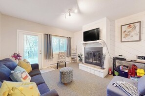 A living room with a sofa, ottoman, fireplace, TV, and a window with curtains. - Ski-in/out condo with a pool, tennis & fast WiFi - Sachem Trail at Okemo (Ludlow)