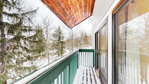 En balkong med ett träpanelat tak, ett grönt räcke och utsikt över snötäckta träd.