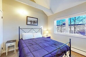 Ein Schlafzimmer mit Bett, Nachttisch, Lampe, einem Bild an der Wand und einem Fenster mit Blick auf Bäume.
