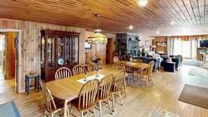 Ein Zimmer mit Holzwänden, einem Esstisch, Stühlen, einer Kristallleuchtre und einem Steinkamin.
