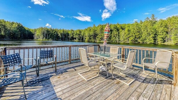 Eine Holzterrasse mit Stühlen und einem Tisch, mit Blick auf einen See, im Hintergrund Bäume.