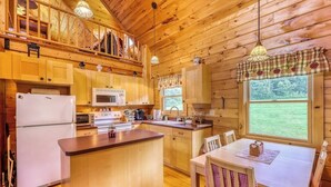 A wooden cabin with a kitchen featuring a refrigerator, microwave, and stove. There is a dining table with chairs and a window with a view of the outdoors.