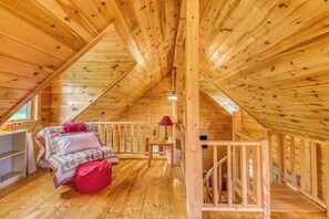 Un interior de madera con techo inclinado, una cama con almohadas, una mesita con una lámpara y una escalera.