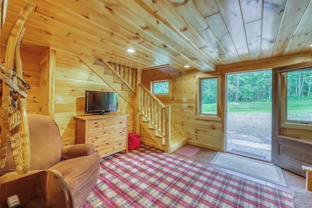 A cabin interior with a plaid carpet, wooden walls, a TV on a dresser, a chair, and a staircase.
