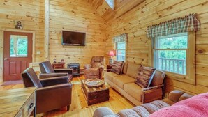 A log cabin interior with a wooden floor, a television mounted on the wall, a fireplace, and a large window with a view of the outdoors.
