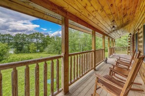 Un porche de madera con barandilla, sillas de madera y vistas a un paisaje verde.